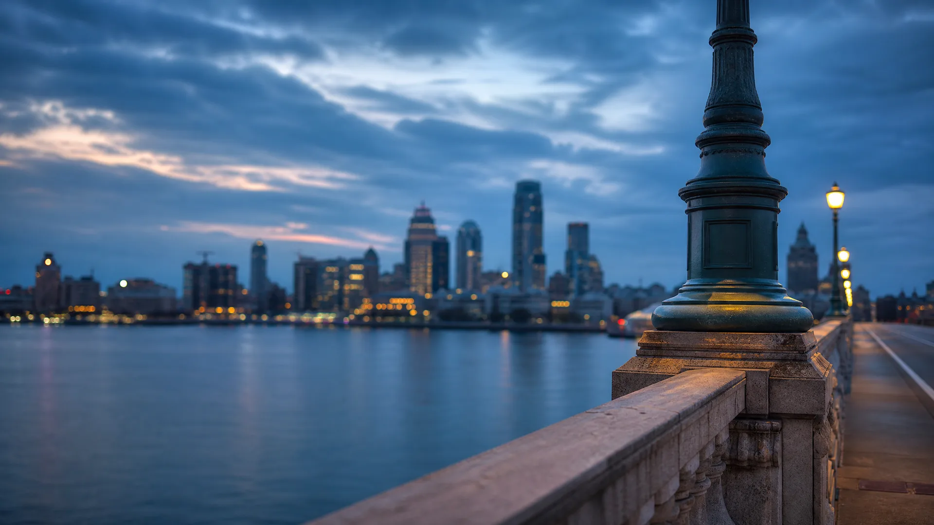A stone bridge balustrade with an ornate lamp post base in the foreground, overlooking water and a blurred city skyline with lit windows under a cloudy evening sky.