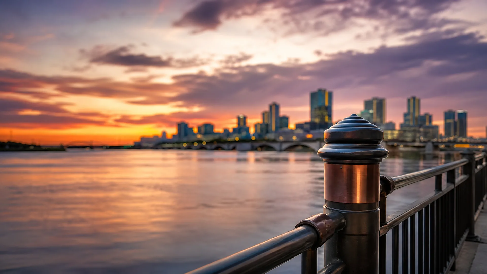 A metal railing post in sharp focus along a waterfront promenade at sunset, with a blurred city skyline and bridge across calm water under purple and orange clouds.