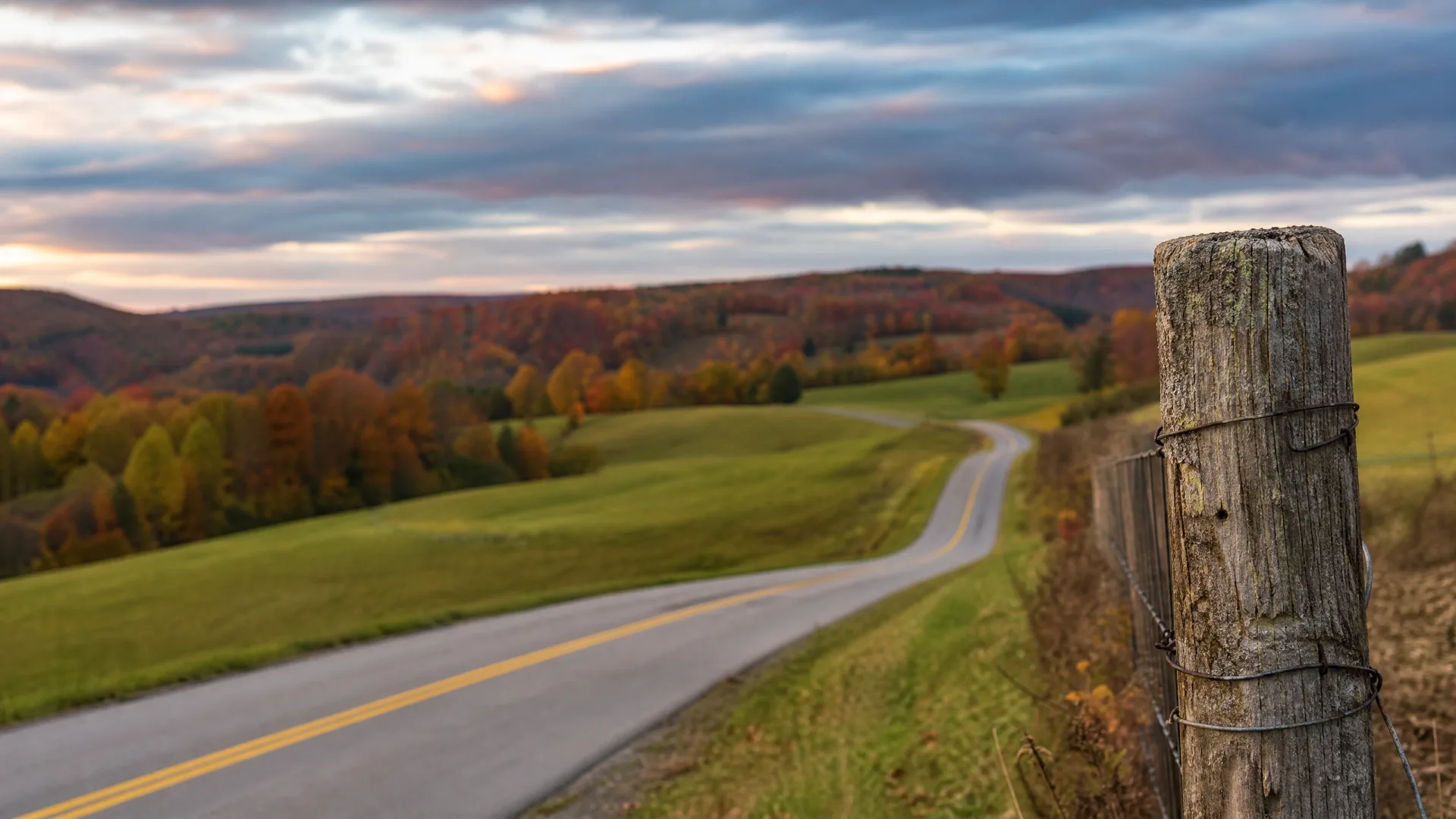 A two-lane road with yellow center lines curves through green fields and autumn-colored hills under a cloudy sky, with a weathered wooden fence post wrapped in wire in sharp focus at the right edge.
