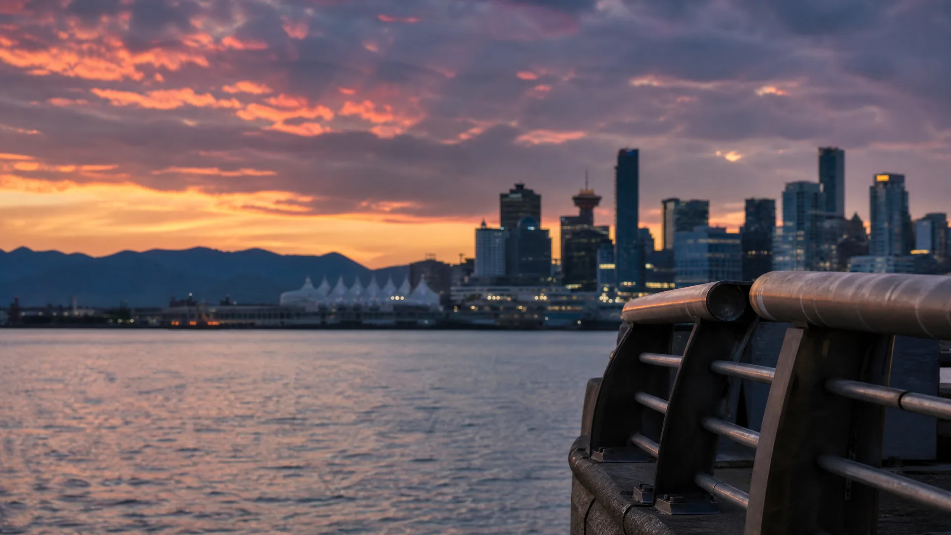 A city skyline of high-rise towers across water at sunset, with distant mountains and an orange and purple cloudy sky, seen past a blurred metal railing in the right foreground.